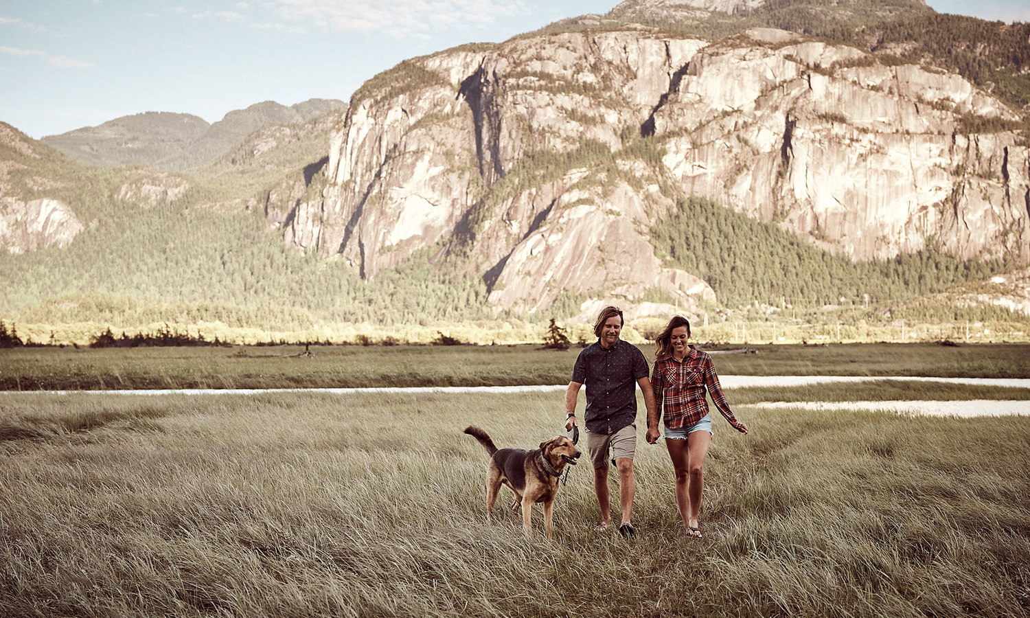 Couple walking with dog in a grassy field with mountains in the background